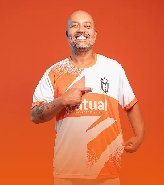 Homem sorridente com os braços cruzados, vestindo uma camiseta cinza escura, contra um fundo laranja.
