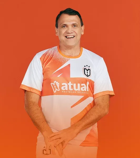Homem sorrindo, vestindo uma camisa laranja, em pé na frente de um carro preto.