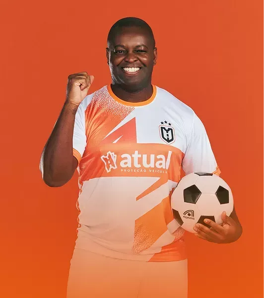 Homem sorridente, vestindo uma camisa polo laranja e com os braços cruzados, está parado ao lado de um carro.