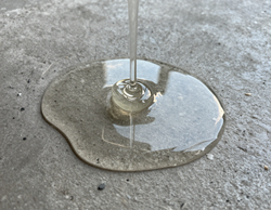 Liquido trasparente che si accumula su una superficie di cemento ruvido attorno a un piccolo elemento metallico centrale.