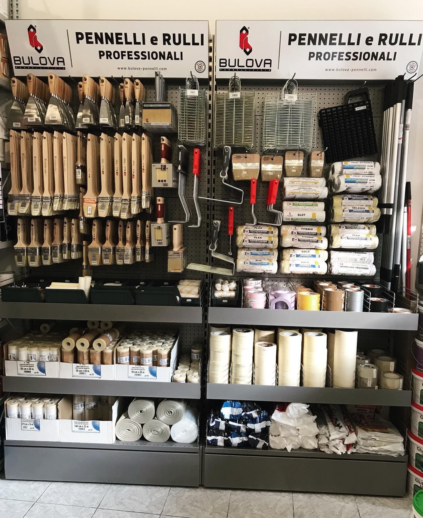 In vetrina, sotto le insegne Pennelli e Rulli, sugli scaffali del negozio sono esposti attrezzi per la pittura, tra cui rulli, pennelli, vassoi e barattoli di vernice.