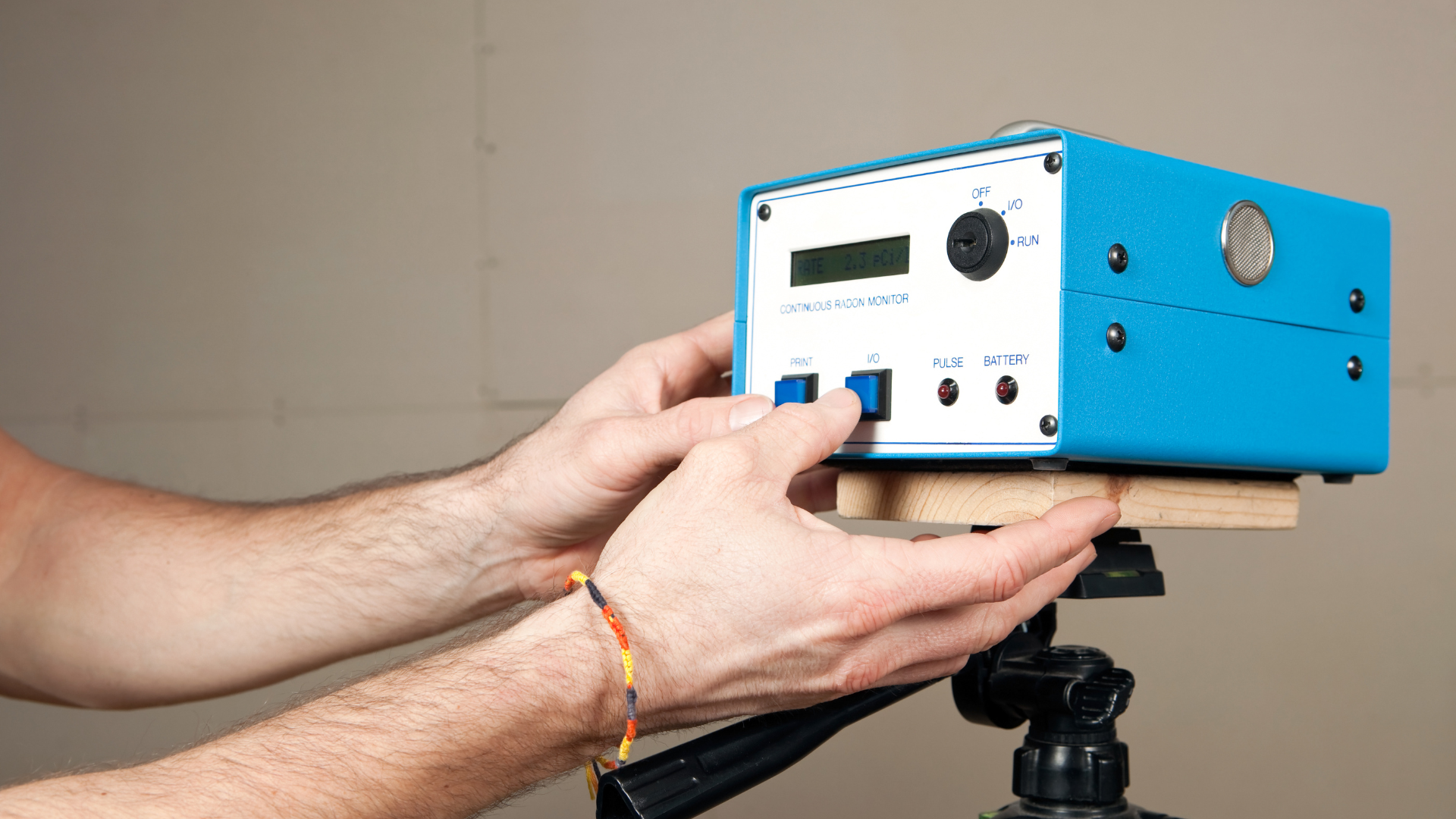 Hands adjusting a blue electronic device mounted on a tripod, with a beige wall background