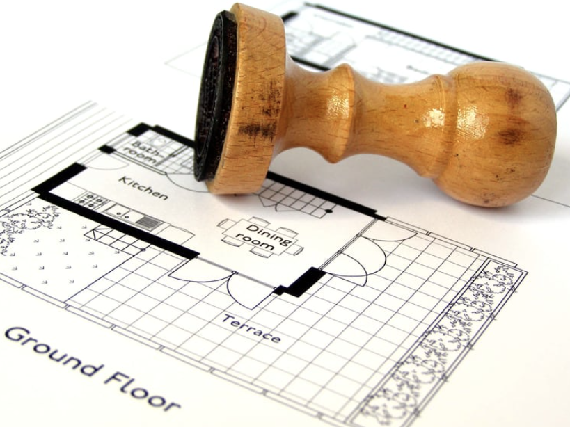 A wooden stamp rests on an architectural floor plan showing a kitchen, dining room, and terrace.