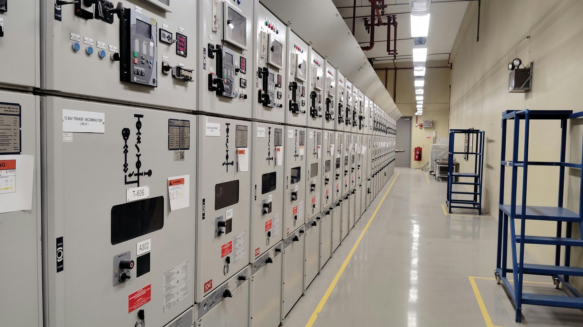 Industrial electrical control room with rows of gray panels and a clear walkway under bright lights