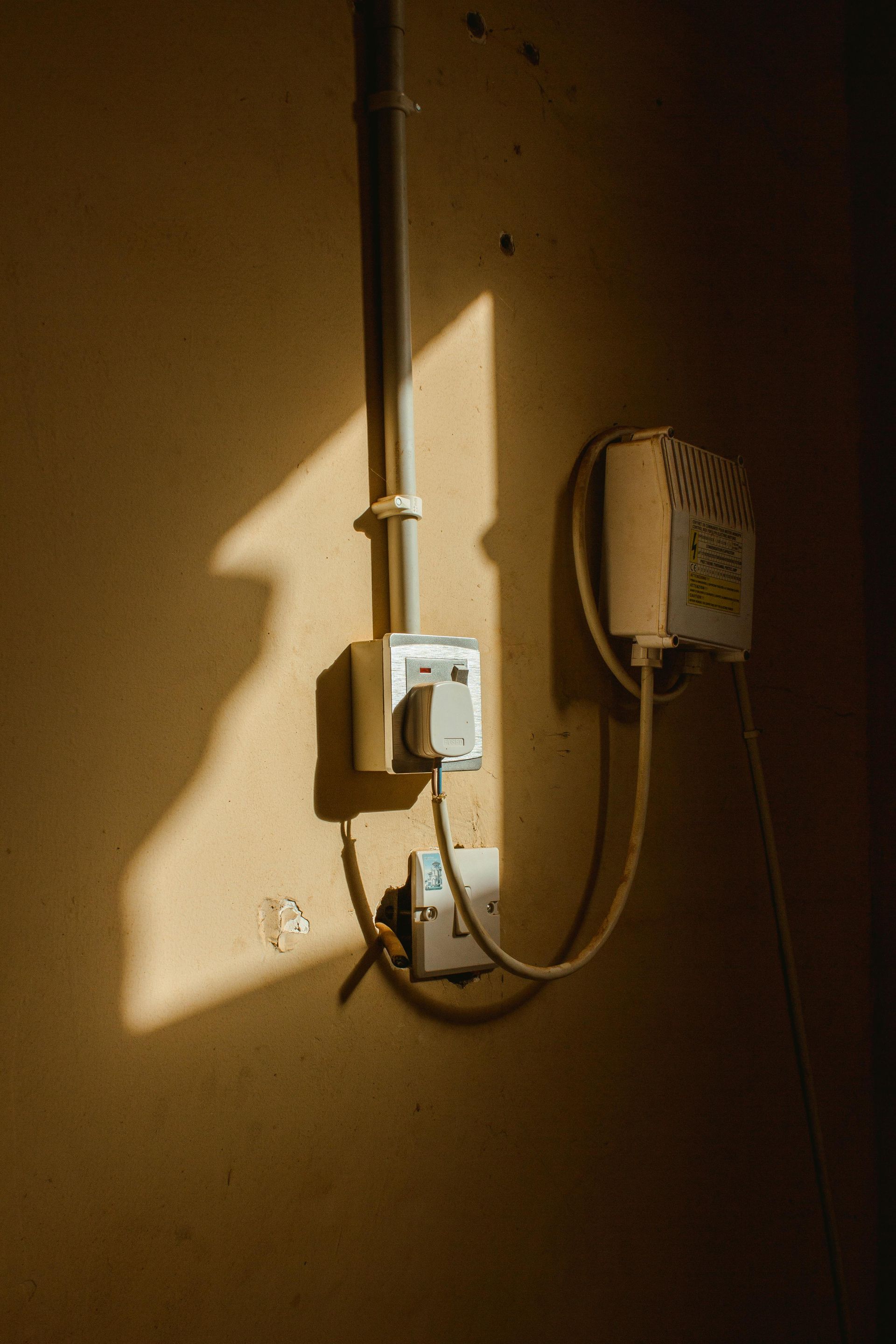 A vertical wall with a conduit, a power outlet, a light switch, and a small white heater plugged into the socket.