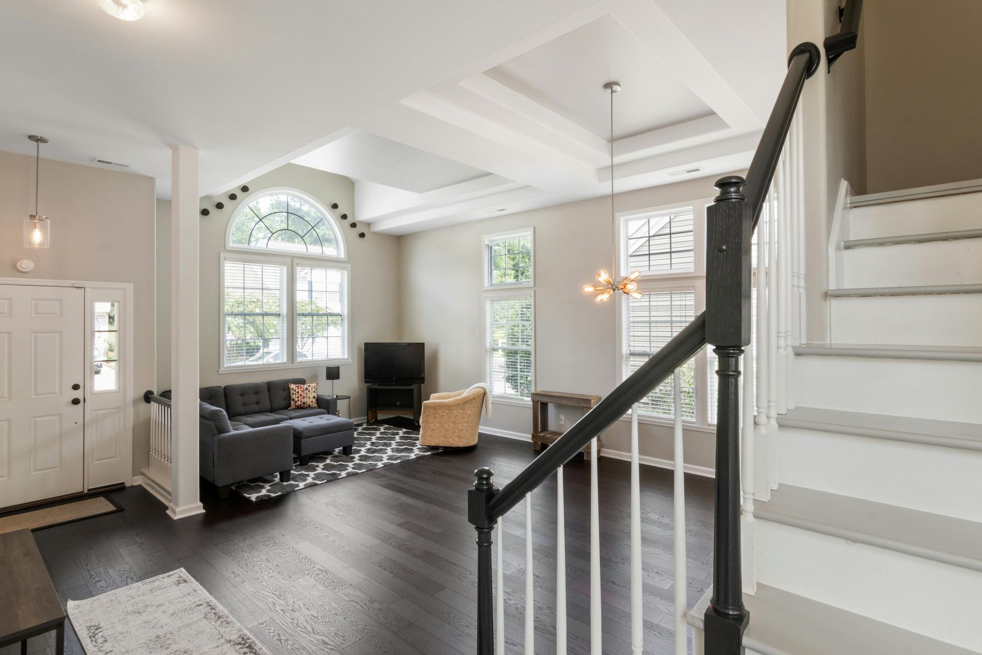 An open-concept living area with dark wood floors, a grey sofa, a television, a tan armchair, and a white staircase.