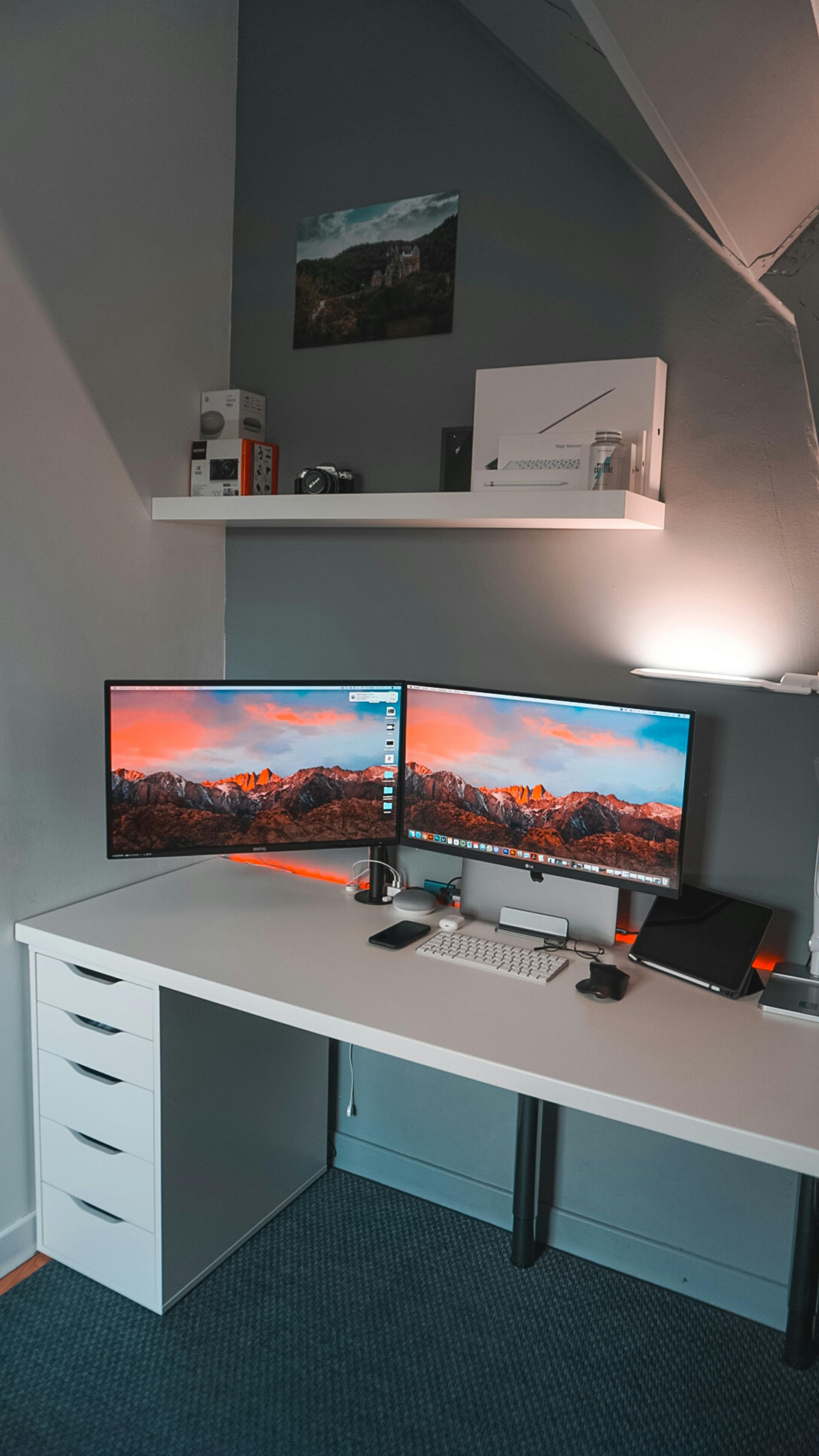 A modern home office with a dual-monitor setup on a white desk, a storage cabinet, and a floating shelf on a gray wall.
