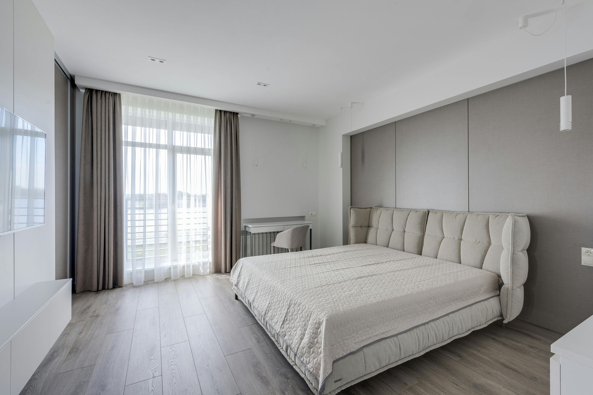 A modern, minimalist bedroom with light wood floors, a plush beige bed, and large windows with floor-length curtains.