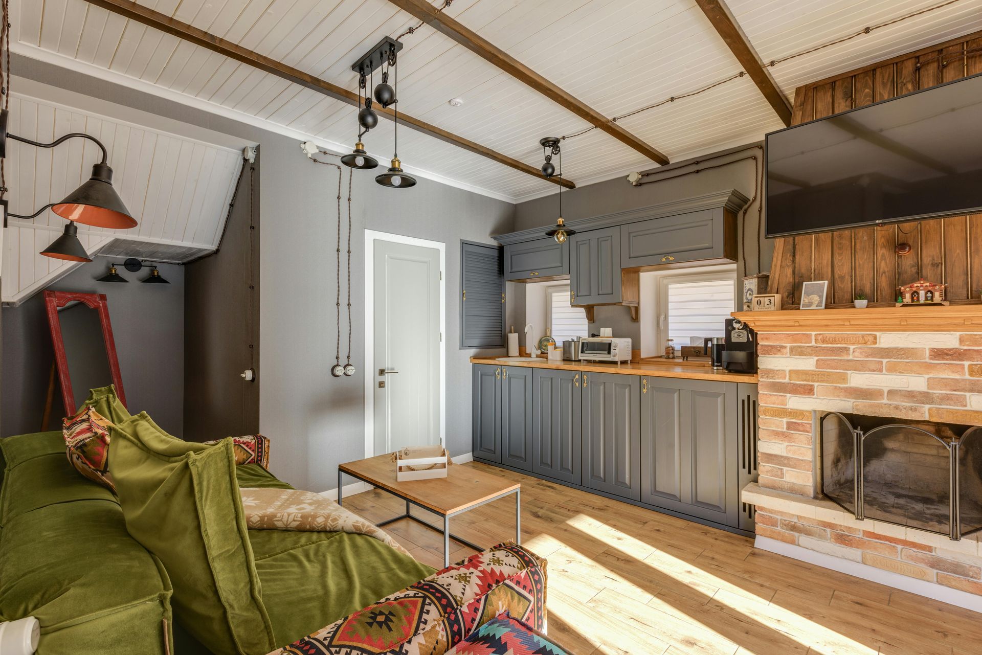 A cozy living room with a green sofa, wooden coffee table, grey kitchen cabinets, and a brick fireplace.