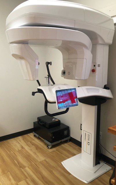 Dental CT scanner, white, in a medical office setting. Shows a large device with a patient headrest and a touch screen display.