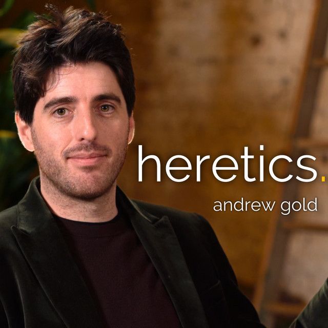 A man in a black jacket is sitting in front of a sign that says heretics