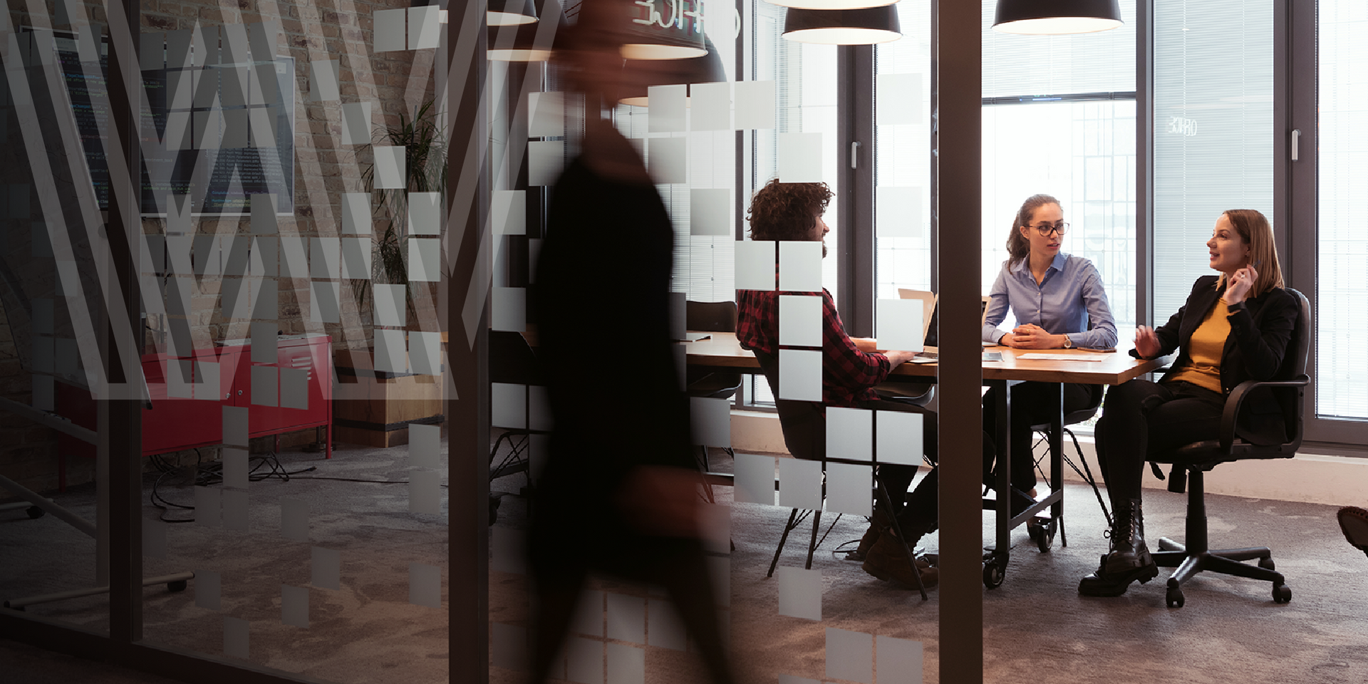 A person walks past a glass-walled conference room where three people sit at a table.