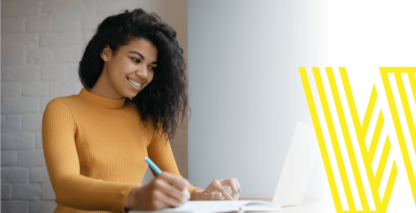 Woman in yellow top writing at a desk, smiling at laptop, near white and yellow logo.