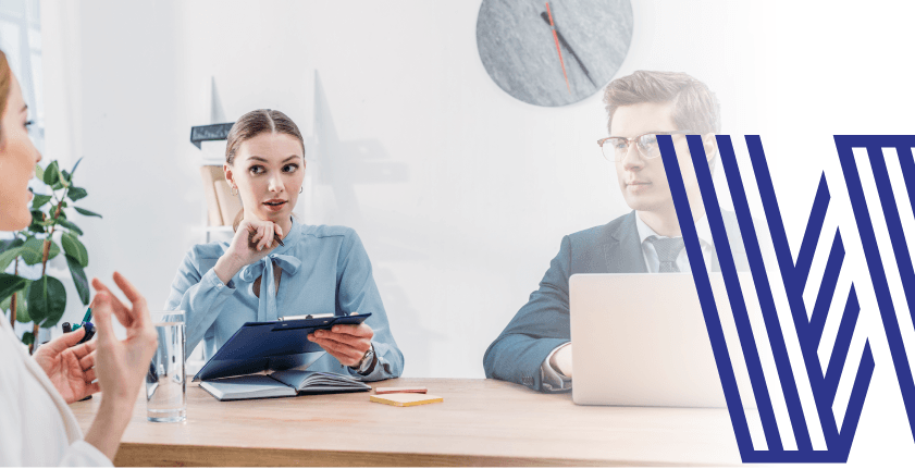Business people in meeting; woman holds clipboard, man uses laptop, clock on wall.