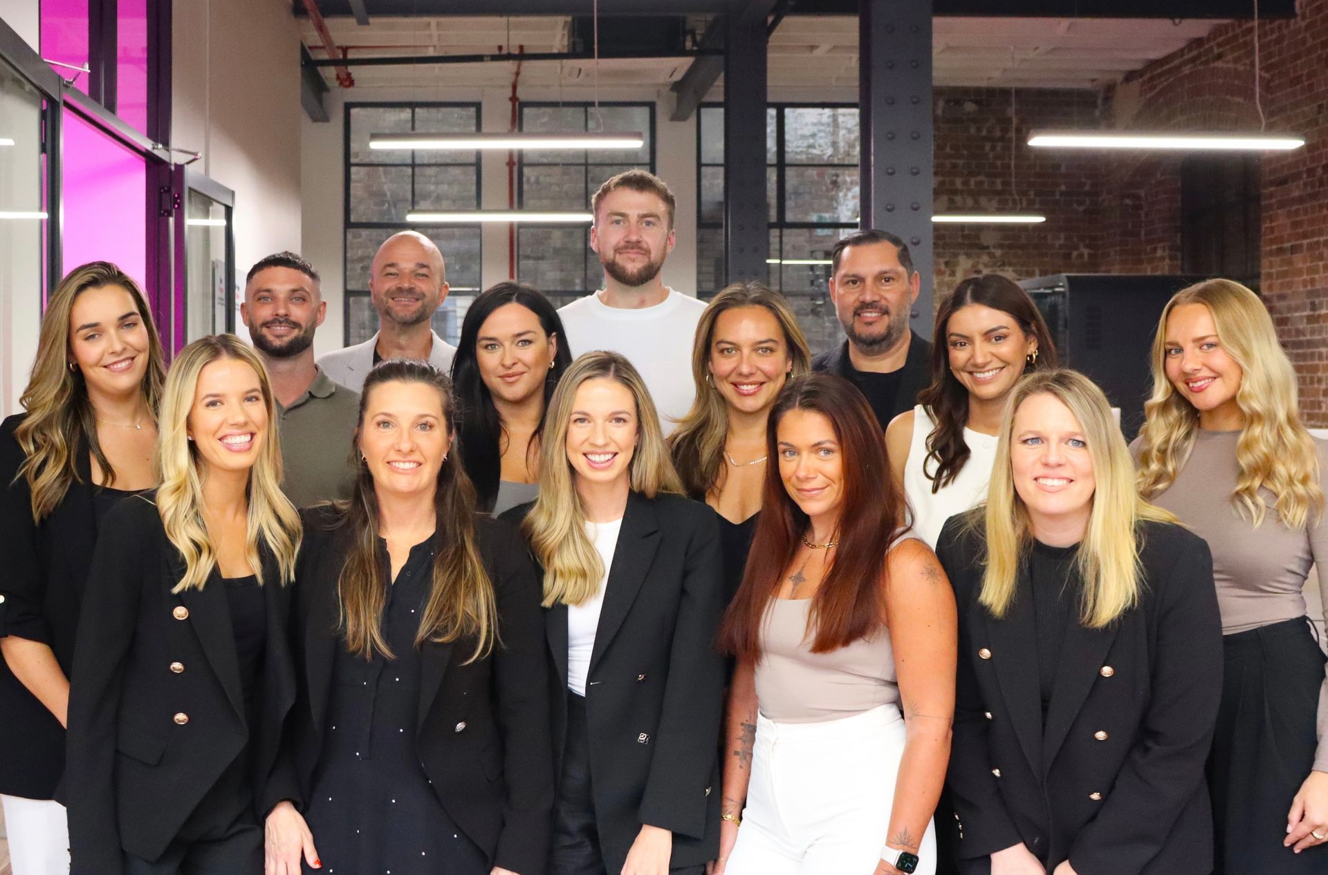 Group of people, smiling, posing in an office. Mostly wearing black blazers. Brick wall backdrop.