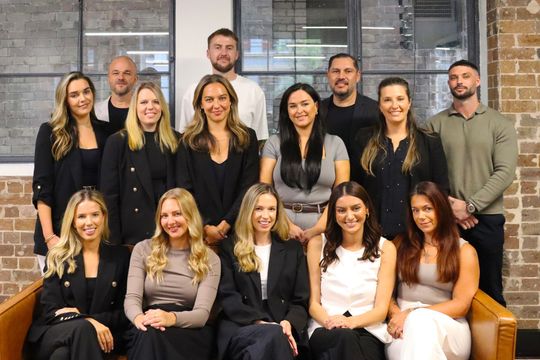 Group photo: 15 people in an office setting. Some are smiling, wearing business attire, and sitting on a brown couch.