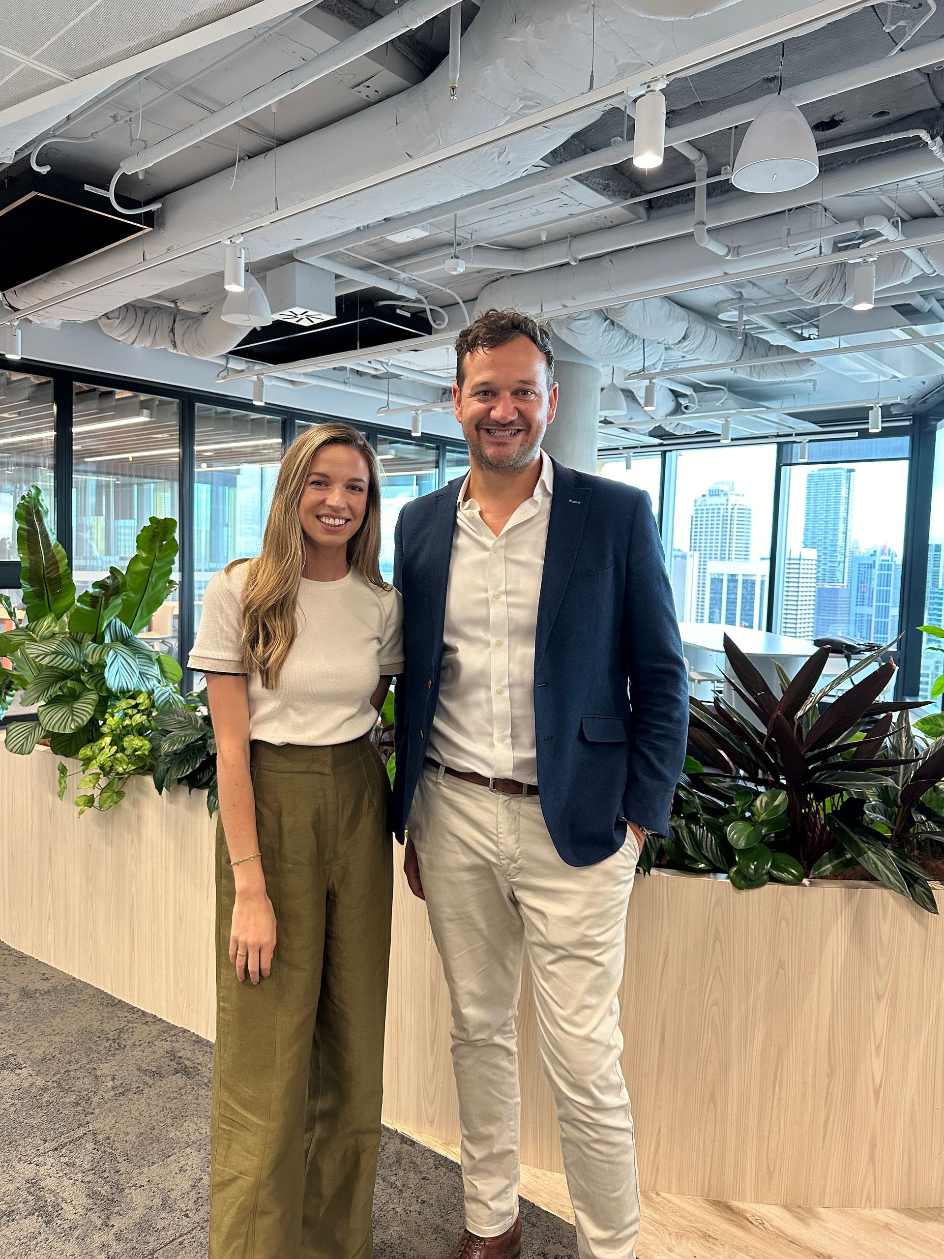 Woman and man posing together, office setting, surrounded by plants. Man in blue blazer, woman in white top.