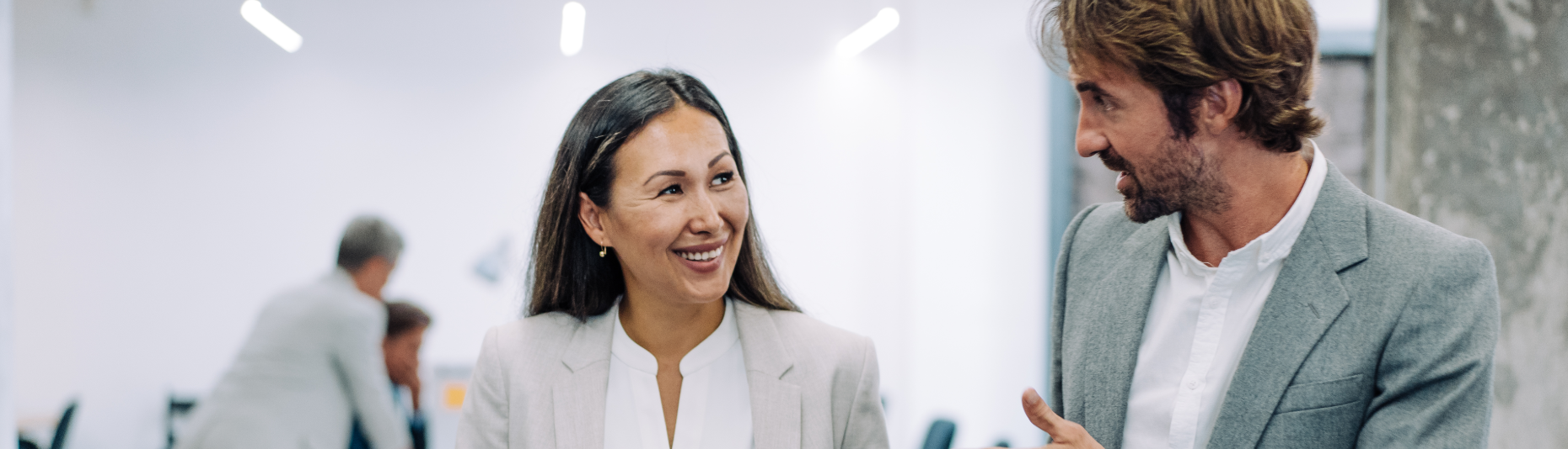 Two people in business attire walking and talking in an office. They are smiling.