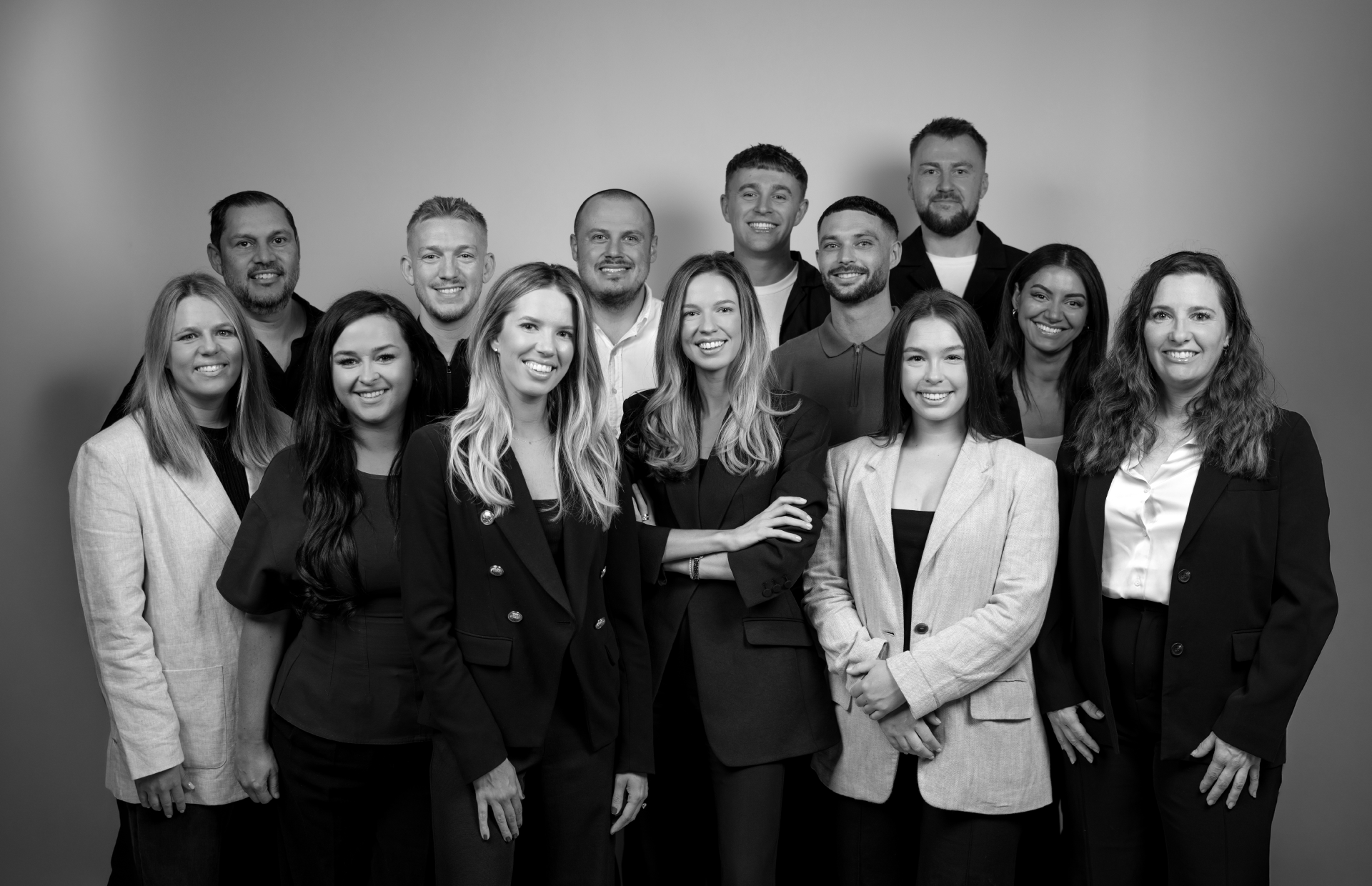 Group of people, smiling, posing in an office. Mostly wearing black blazers. Brick wall backdrop.