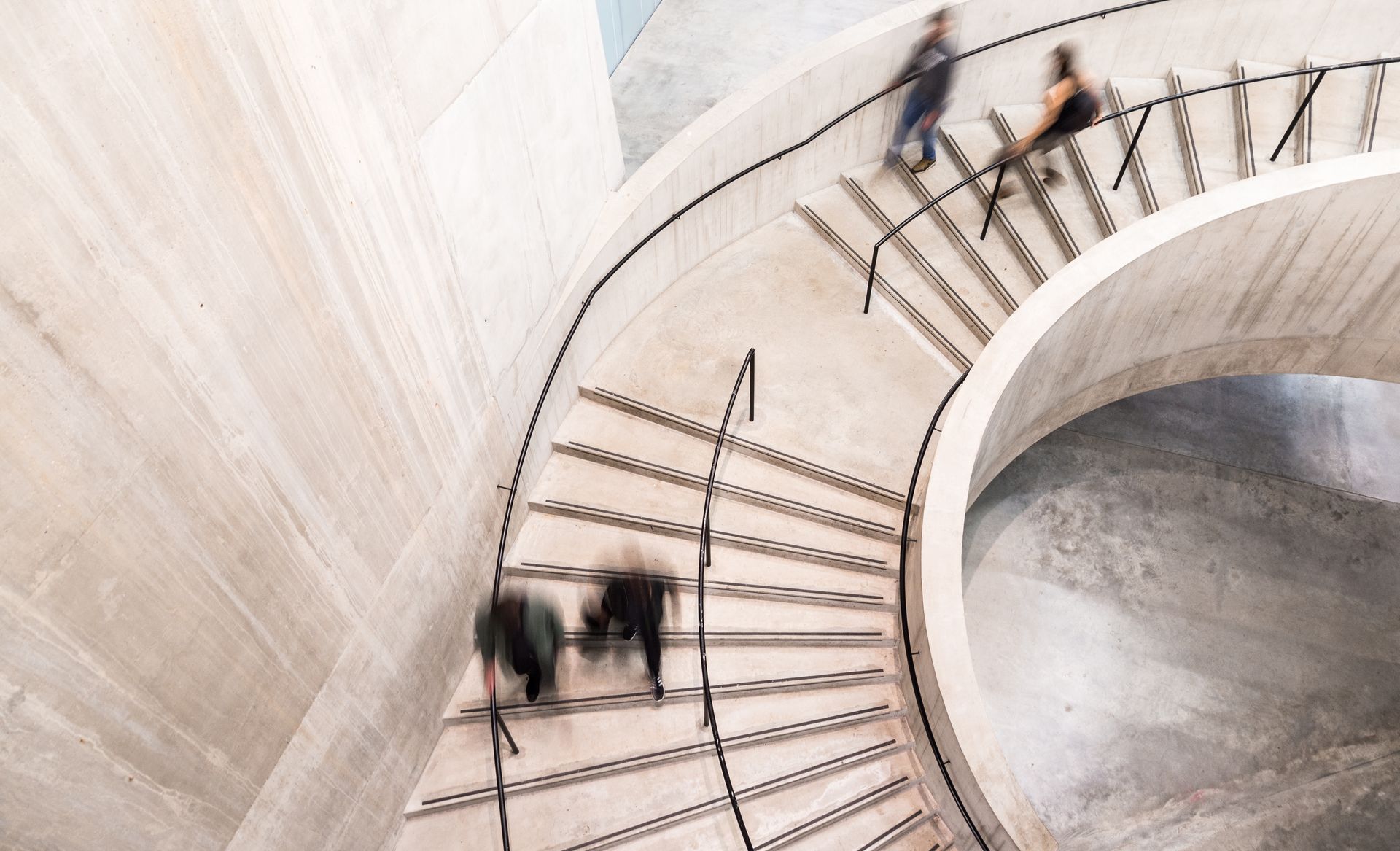 Spiral staircase with blurred figures ascending and descending. Concrete walls and metal railings.