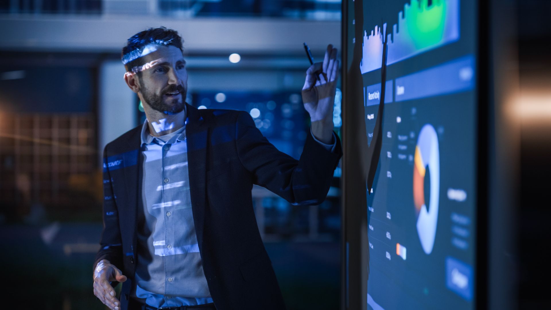 Man in suit points to a data screen with charts and graphs. Night setting with blue lighting.