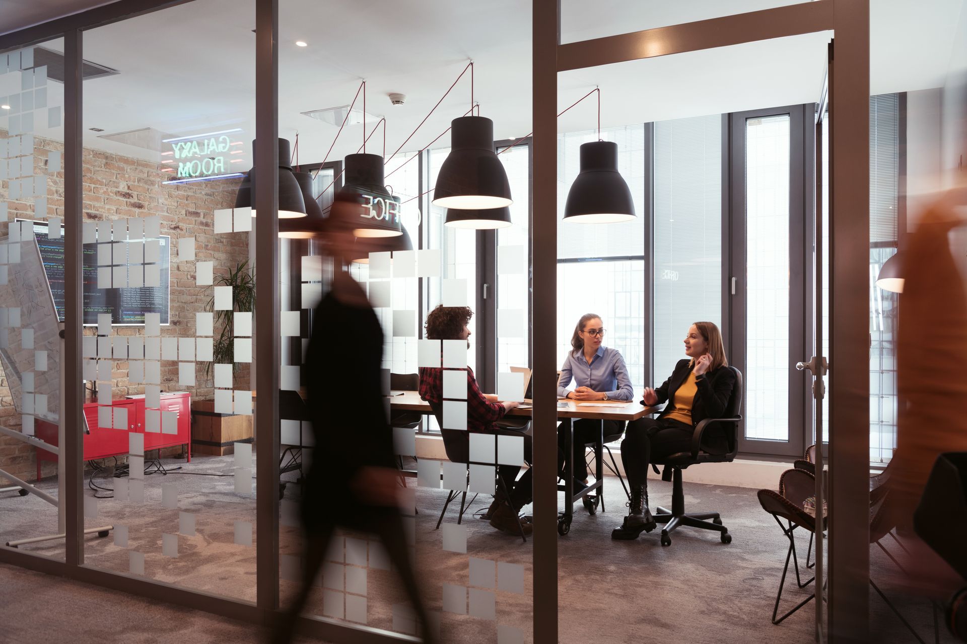 Blurred person walks past a glass-walled conference room where a meeting is in progress.