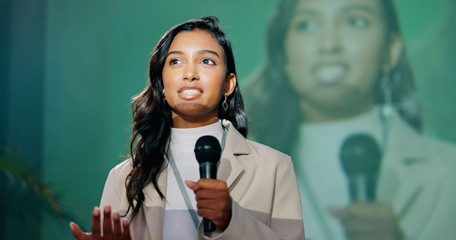 Woman speaking into a microphone, gesturing with her hands. Background shows her image on a screen, with green hues.