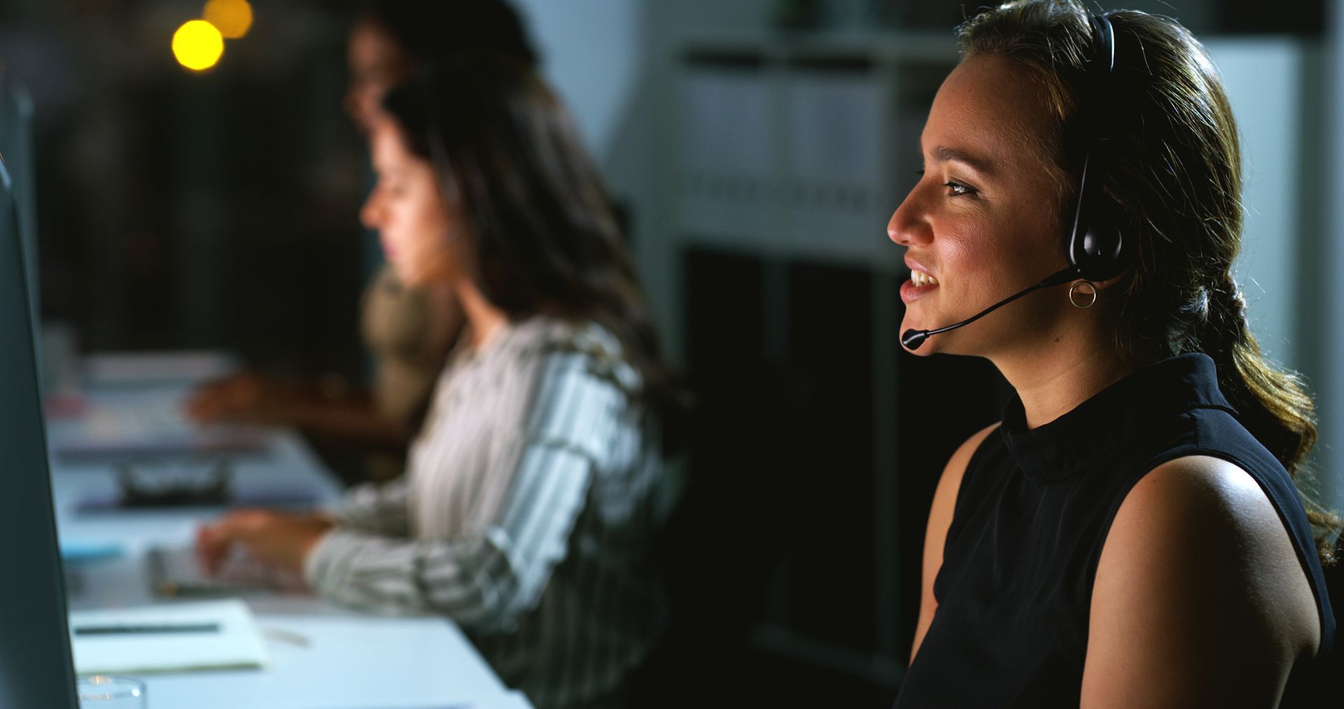 Woman with headset smiles while working at a computer in a dimly lit office.