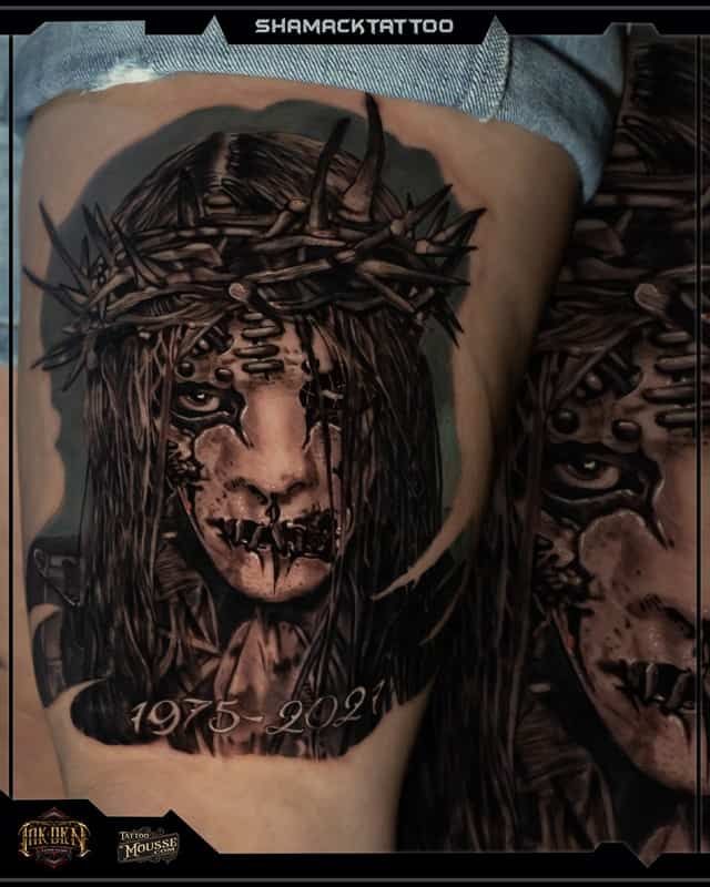 Black-and-gray thigh tattoos of a crowned, bleeding face and a dark portrait with “1976–2021” text