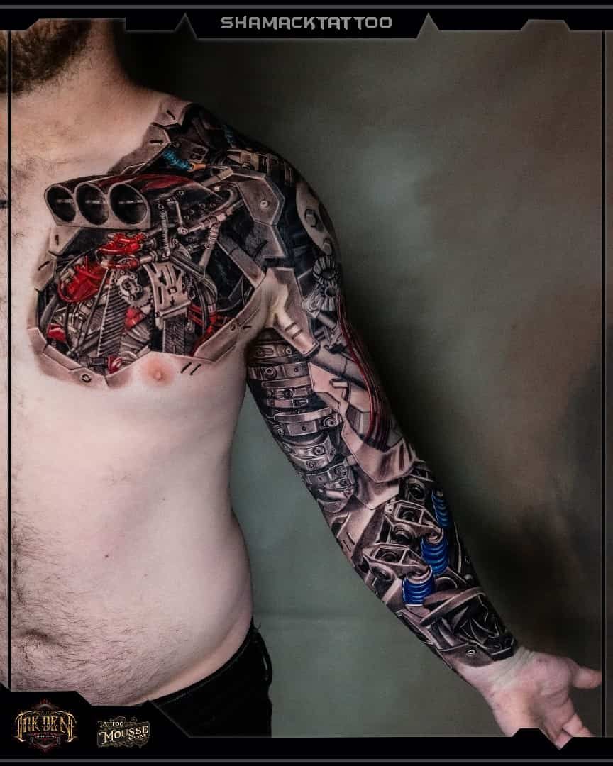 Person with a large biomechanical tattoo covering chest and arm, standing against a plain wall.