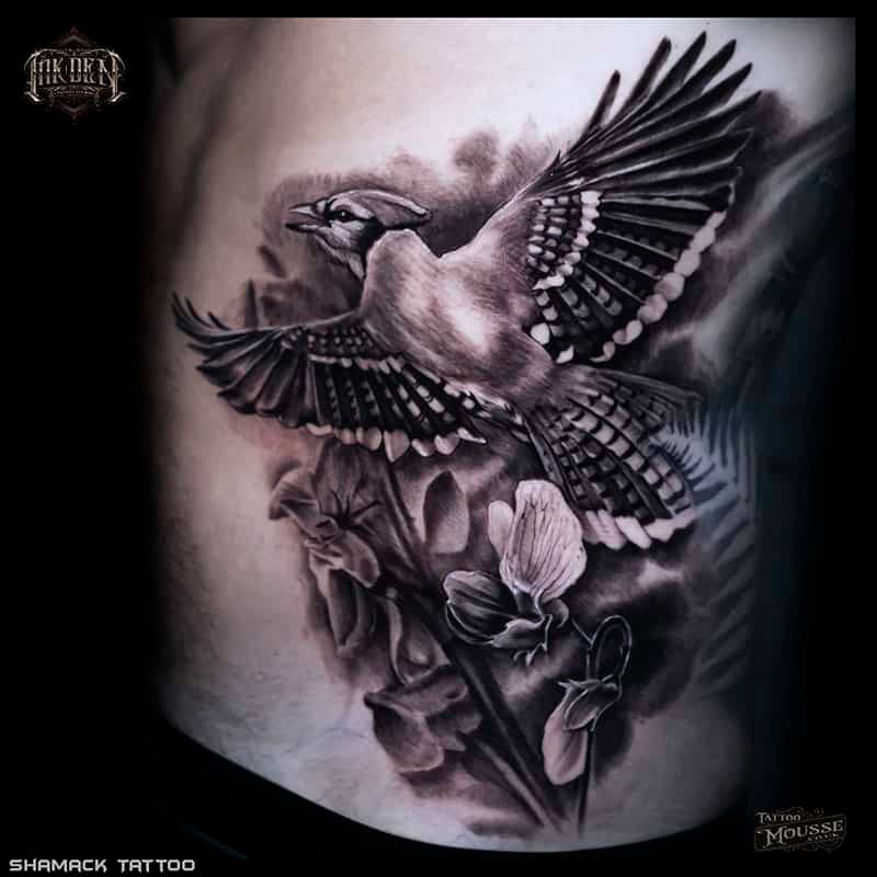 Black and gray tattoo of a flying bird above flowers and leaves on skin