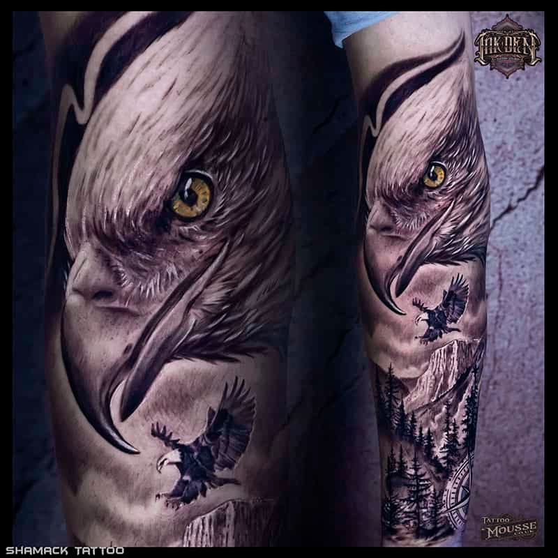 Close-up of a realistic owl tattoo on skin in gray and black ink, with yellow eyes and a forest scene.
