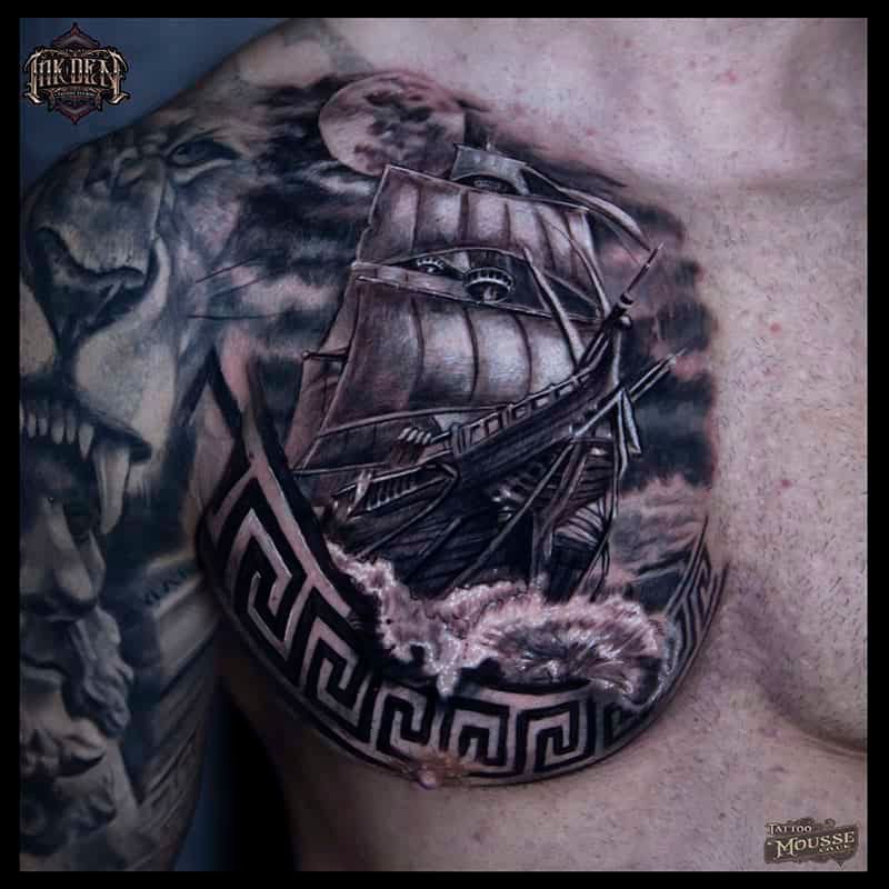 Black and gray chest tattoo of a sailing ship over a lion and Greek key pattern