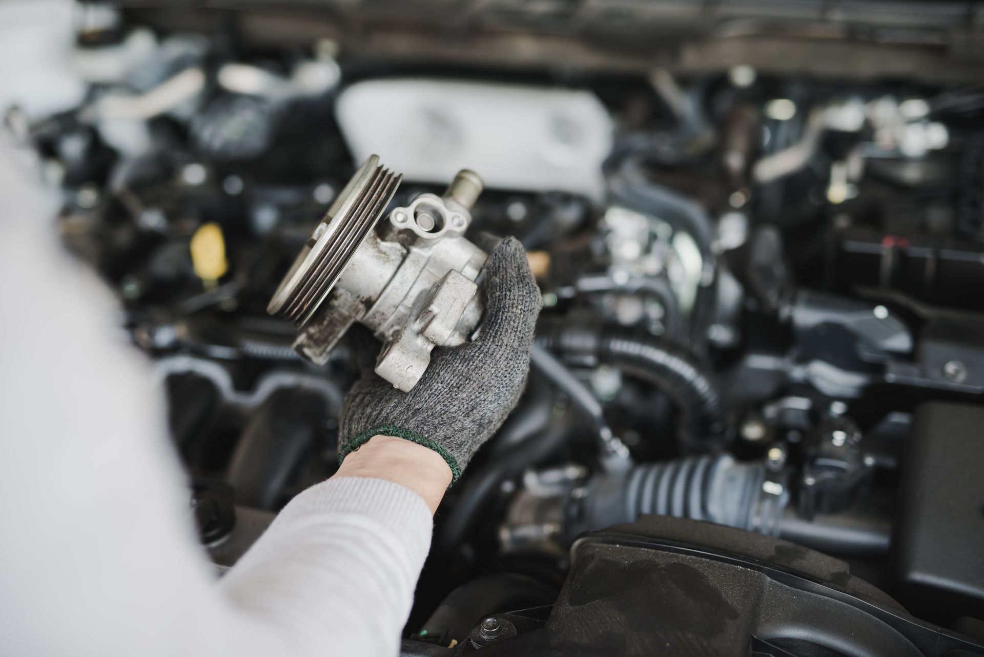 A gloved hand holding a metallic automotive water pump in front of an open car engine.