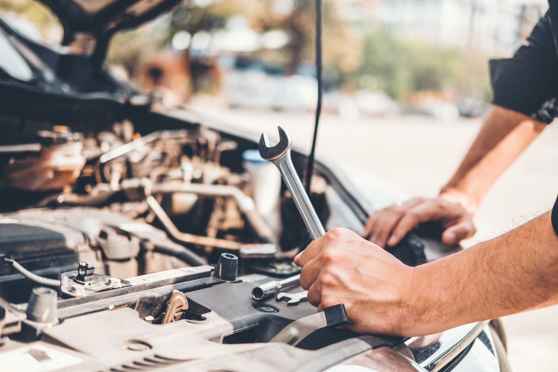 A close-up of a person's hands holding a wrench over an open car engine, with the hood up, during a repair.