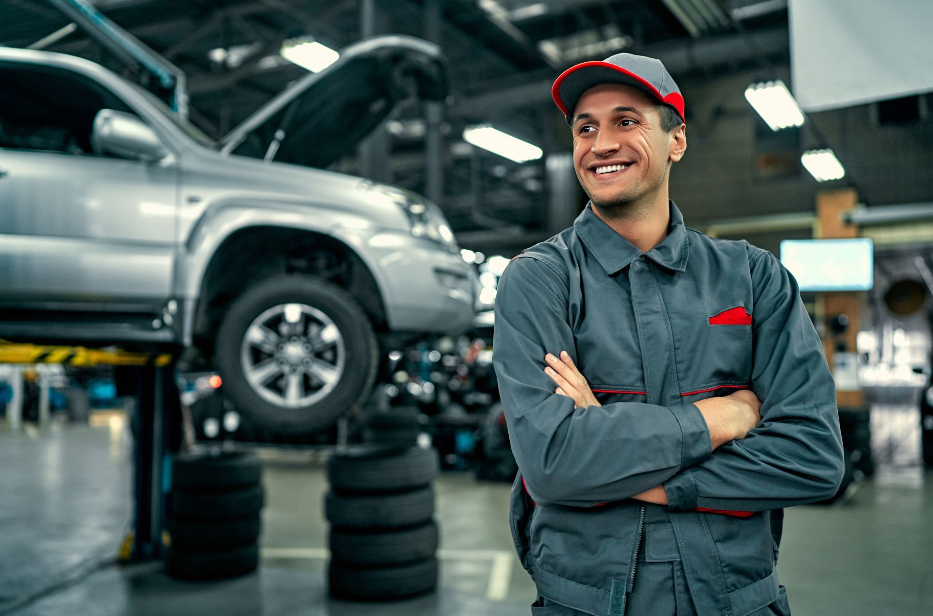 A smiling mechanic in a gray uniform stands with arms crossed in an auto repair shop with a car on a lift.