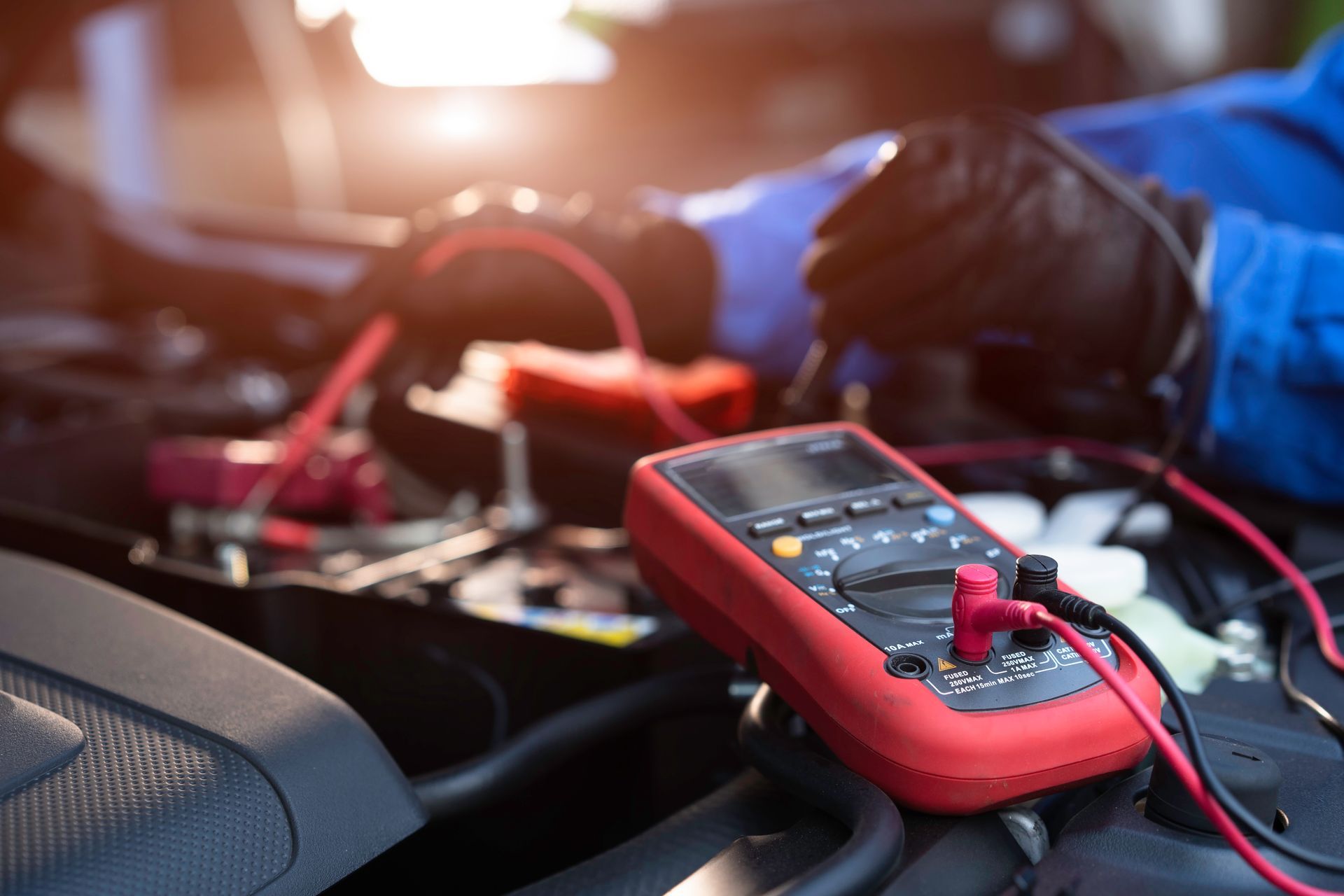 A person wearing black gloves uses a red digital multimeter to test a car battery under bright, sunny conditions.