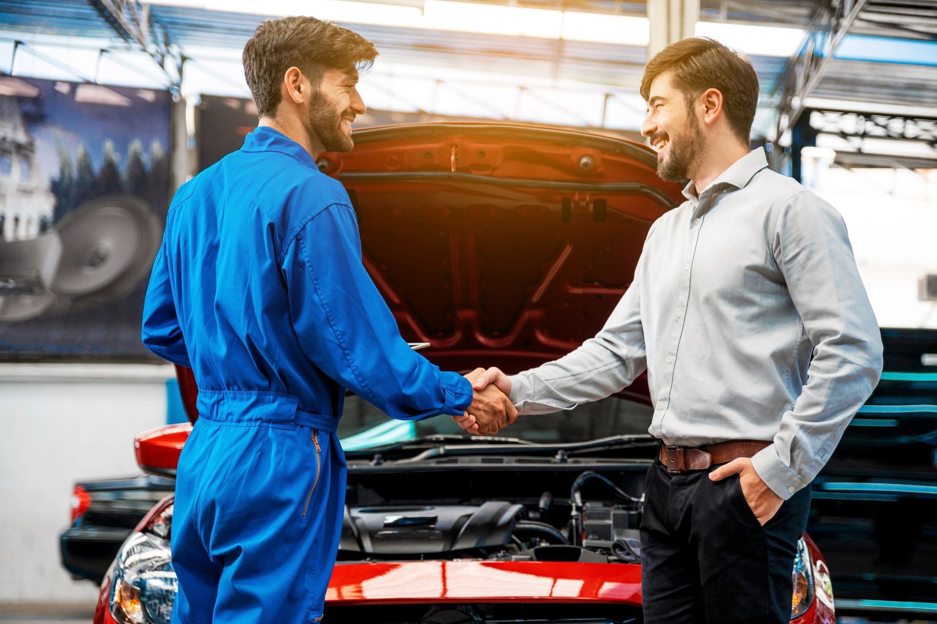 A mechanic in blue coveralls and a customer in a button-down shirt shake hands in front of a car with an open hood.