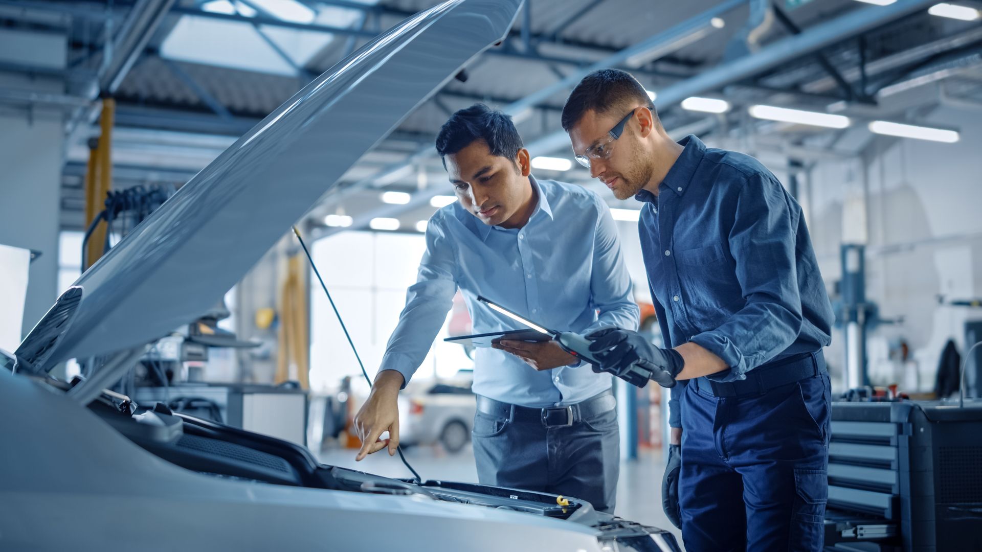 Two professionals examine a car's open engine bay in a brightly lit automotive repair shop.