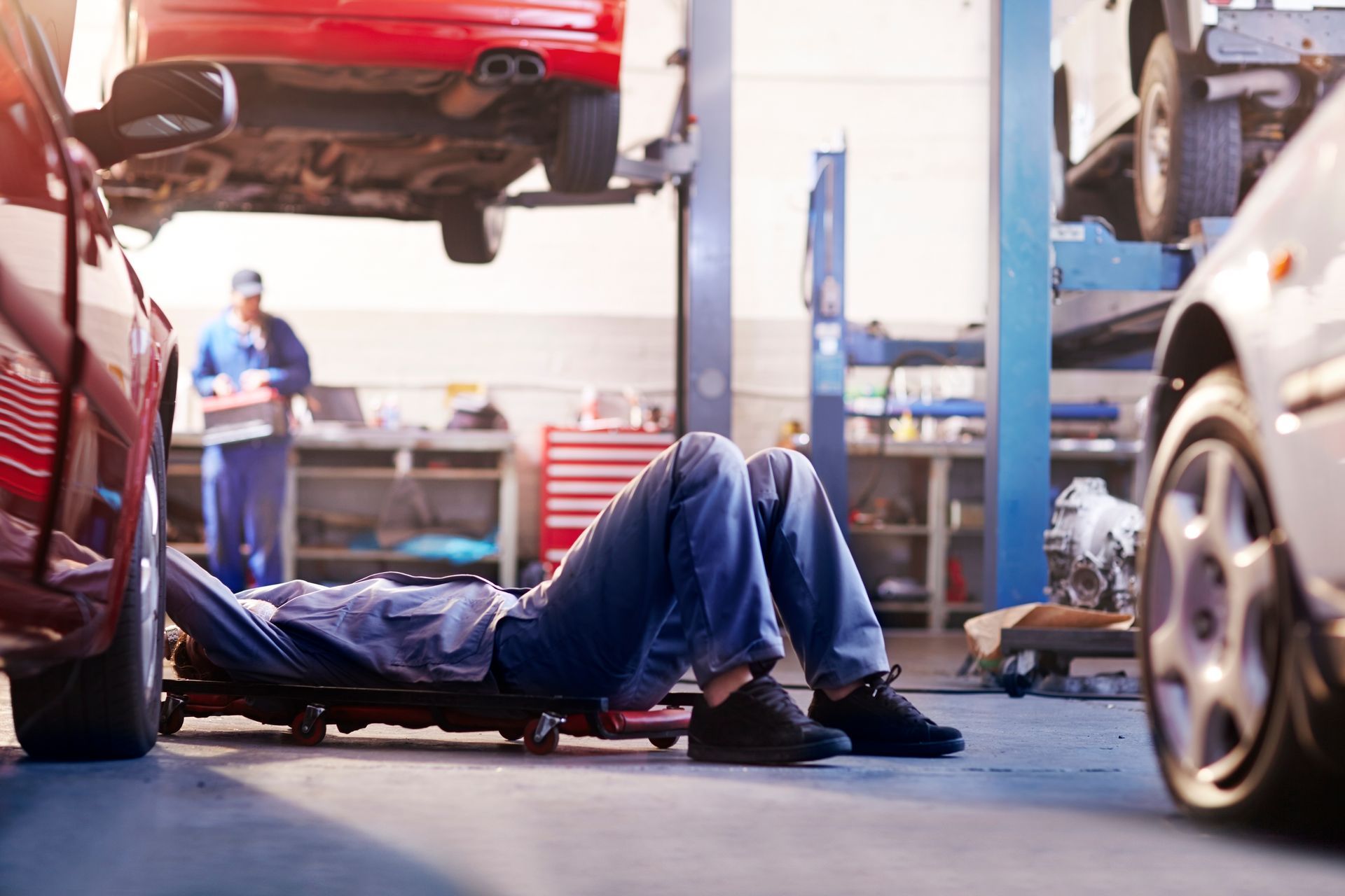 A mechanic lies on a creeper working under a car in an auto shop with another person in the background.