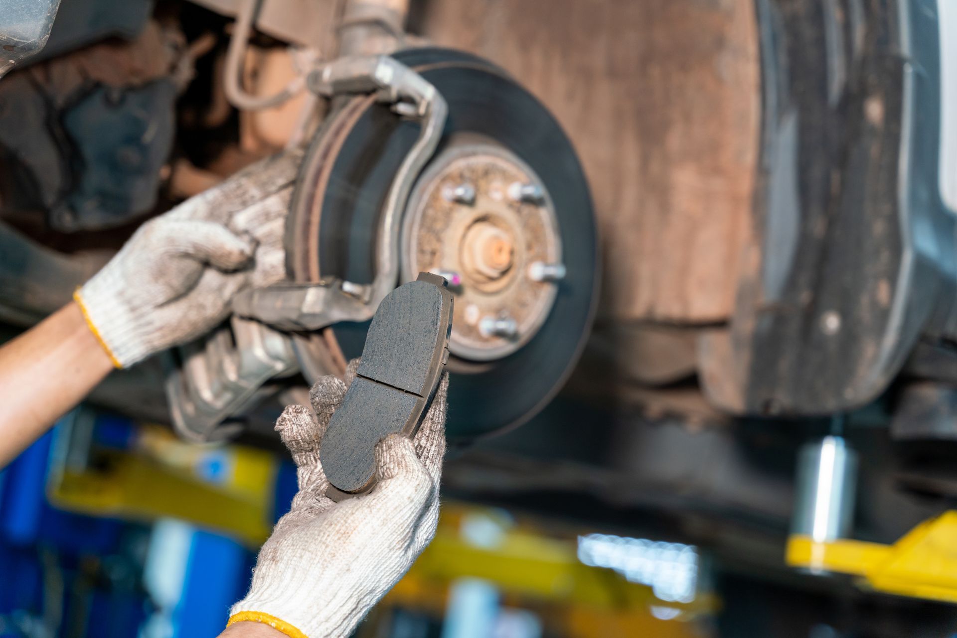 A mechanic wearing gloves holds a new brake pad in front of a car's disc brake assembly in an auto repair shop.