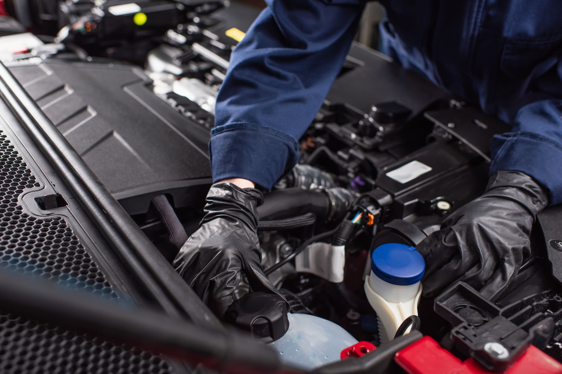 A technician wearing blue coveralls and black gloves performs maintenance on the engine bay of a car.