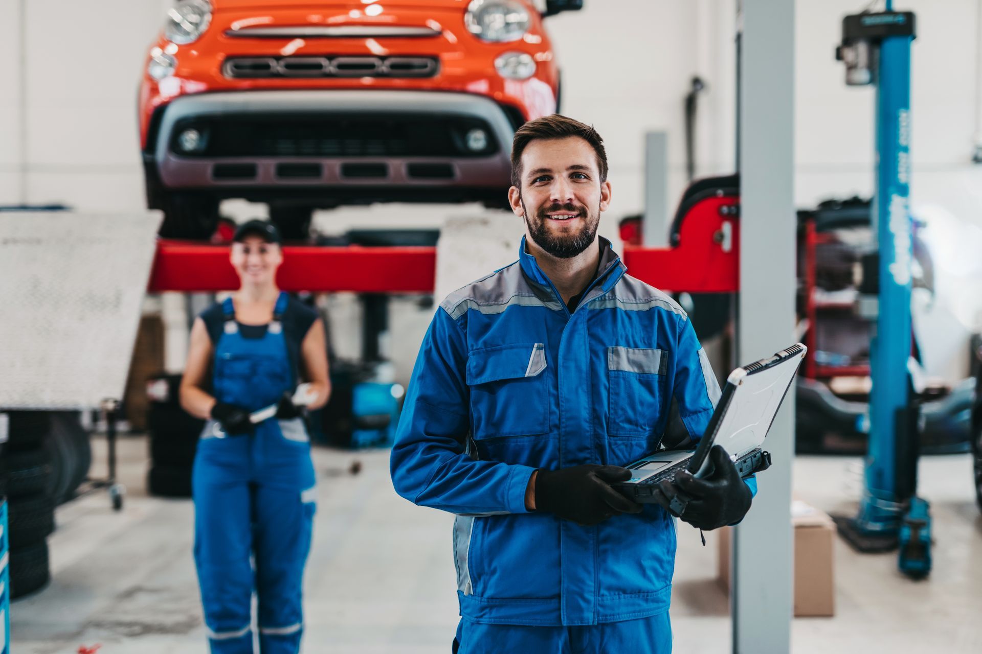 A smiling mechanic in a blue uniform holds a tablet in a garage, with another mechanic and a lifted car in the background.