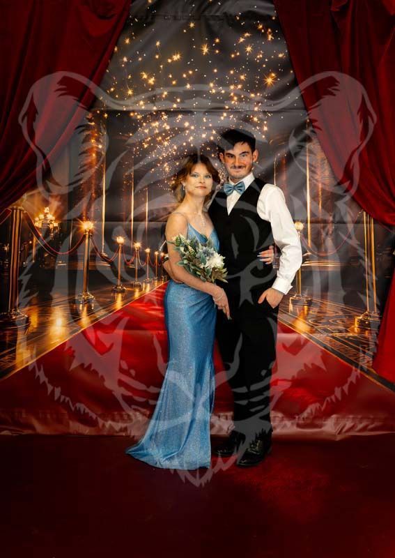 A couple in formal attire stands on a red carpet in front of a backdrop featuring a ballroom and string lights.