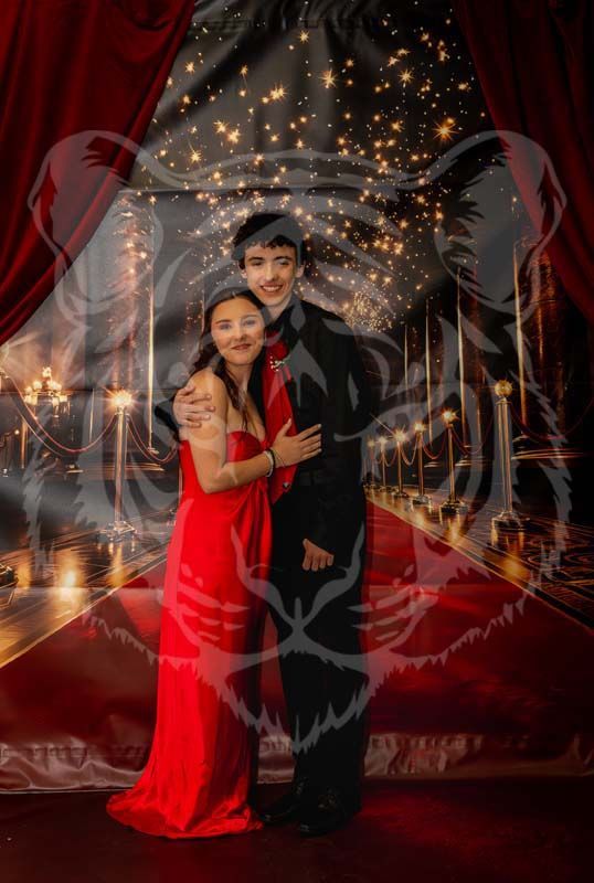A couple in formal attire, one wearing a red gown and one in a black suit, poses on a red carpet backdrop with lights.