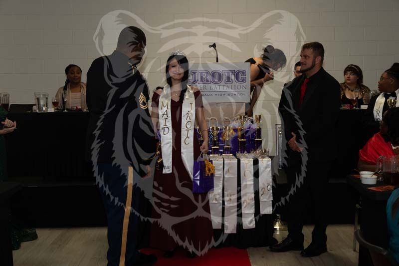 A person wearing a maroon dress and a white sash stands before a table of trophies at a formal event.