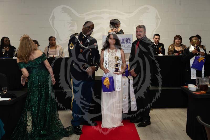 A person in a white gown holds a trophy on a red carpet between two others, surrounded by guests in a formal hall.