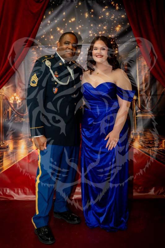 A man in a military dress uniform and a woman in a blue formal gown pose together in front of a red-curtained backdrop.