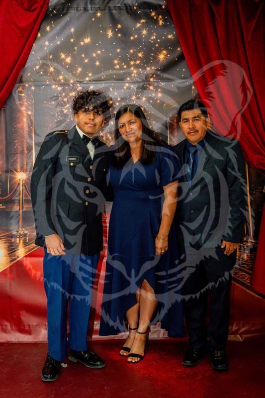 Three people stand smiling in front of a red-curtained backdrop with string lights at a formal event.
