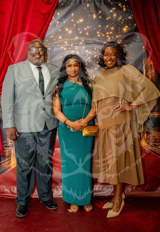 Three people pose for a photo in front of a red curtain and fairy light backdrop, smiling at the camera.